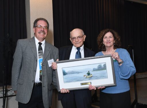 Three formally dressed people standing together for a picture. The man in the middle is holding an award.
