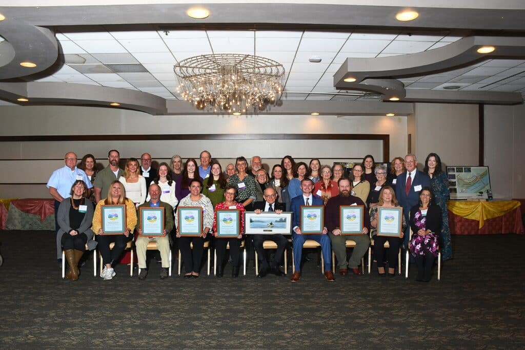 A group photo of men and women. A row of people are sitting with awards with another row of people standing behind them.